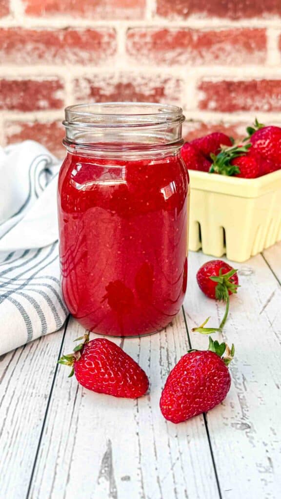 fresh strawberry sauce in a glass with 2 strawberries laying in front and a yellow strawberry basket behind it