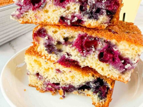 Stacked up blueberry cake squares on a white plate. 3 cake squares high