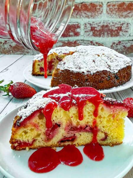 Strawberry Cake slice on white plate being drizzled with strawberry sauce on top