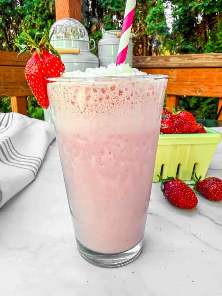 A tall strawberry milk shake topped with whipped cream and a red and white stripped straw. In the background a yellow basket of strawberries
