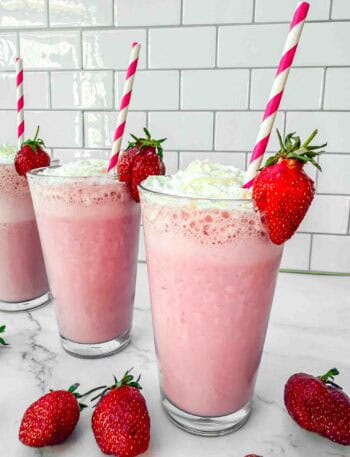 3 glasses of strawberry milkshakes topped with whipping cream, a strawberry on the side and red-white striped straws in the glass