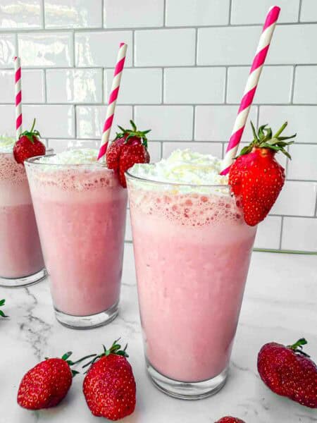 3 glasses of strawberry milkshakes topped with whipping cream, a strawberry on the side and red-white striped straws in the glass