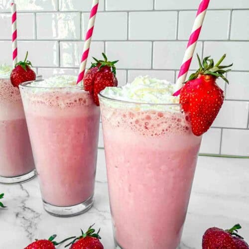 3 glasses of strawberry milkshakes topped with whipping cream, a strawberry on the side and red-white striped straws in the glass