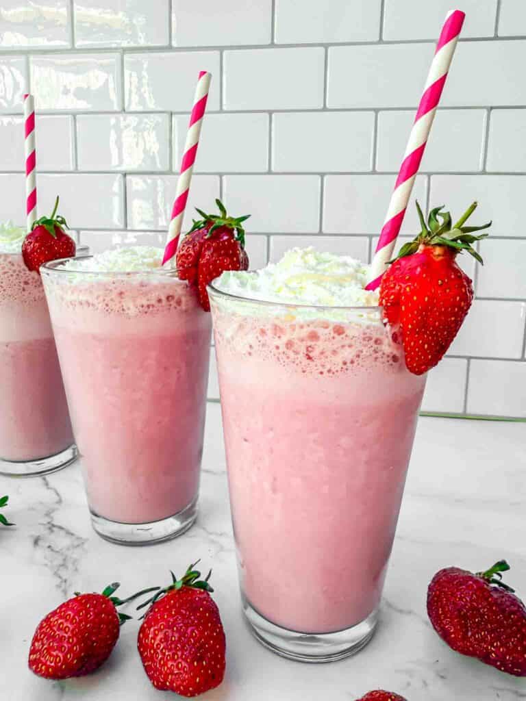 3 glasses of strawberry milkshakes topped with whipping cream, a strawberry on the side and red-white striped straws in the glass